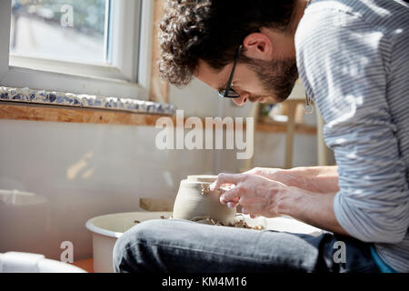 Man seated at a potter's wheel, moulding the wet clay on the spinning turntable, in a pottery studio Stock Photo