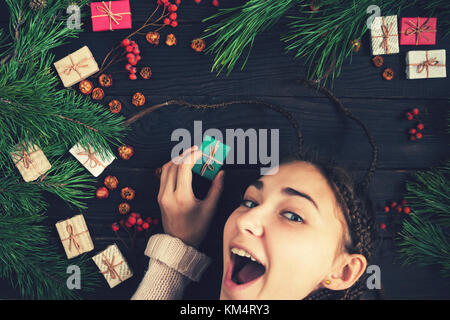 A girl is holding a Christmas gift and smiling on the background of pine branches and a wooden surface Stock Photo