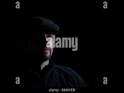 Low-key photo portrait of bearded man in dark cap, half-lit face, pensive pose. Low-key lighting, black background: artistic chiaroscuro effect. Stock Photo