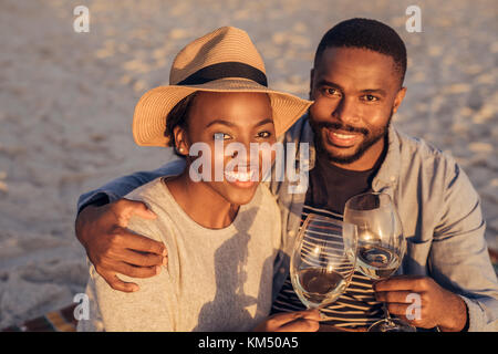 Smiling African couple sitting together at the beach drinking wine  Stock Photo