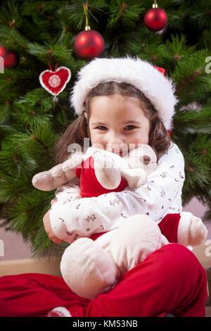 Little child girl hugging her fluffy reindeer toy close to Christmas tree. She is very, very happy Stock Photo