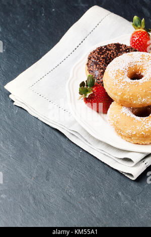 White plate of sugar and chocolate donuts and fresh strawberries served on gray textile napkin over black stone table. See series Stock Photo