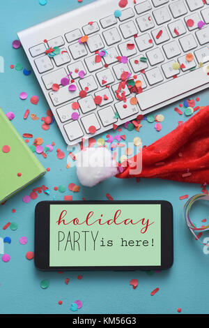 a smartphone with the text holiday party is here in its screen, on an office desk full of confetti, next to a santa hat, a computer keyboard and a not Stock Photo