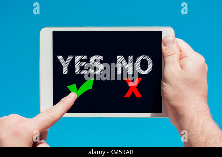 Man holding a tablet device with 'yes no' icons as decision concept and touching the screen with a finger with blue sky in background. Stock Photo