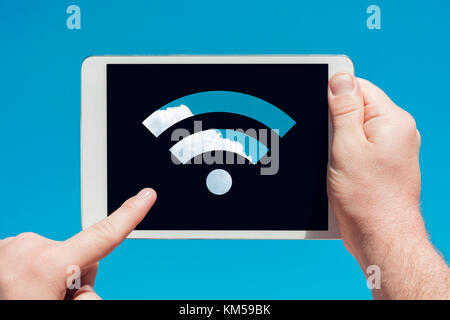 Man holding a tablet device showing wifi sign and touching the screen with a finger with blue sky in background. Stock Photo