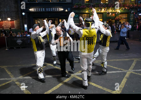 Morris dancers from the Westminster Morris Men, performing outside in Borough Market one Summer evening at people out for an evening drink watch on in London, United Kingdom. The Westminster Morris Men perform with sticks. Morris dance is a form of English folk dance usually accompanied by music. It is based on rhythmic stepping and the execution of choreographed figures by a group of dancers, usually wearing bell pads on their shins. The earliest known and surviving English written mention of Morris dance is dated to 1448. Stock Photo