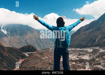 Standing young woman with backpack and raised up arms on the hill and looking on mountains. Landscape with happy girl, mountains, blue sky with clouds Stock Photo