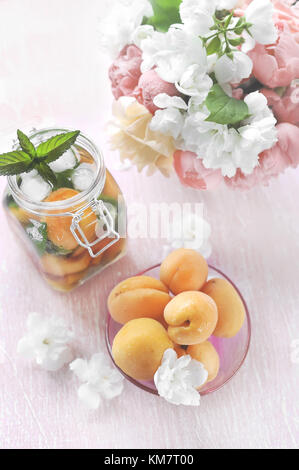 Ripe yellow apricots in a transparent vase with a detox drink made from mint and dried apricots in the background with bouquet of peonies. Stock Photo