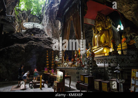 Kanchanaburi, Thailand - July 12, 2017: Three visitors are standing in front of a Buddha statue at the Wat Ban Tham cave temple, which entrance  is form like a dragon's mouth.