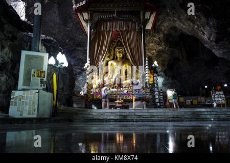 Kanchanaburi, Thailand - July 12, 2017: Two visitors are standing in front of a Buddha statue at the Wat Ban Tham cave temple, which entrance  is form like a dragon's mouth.