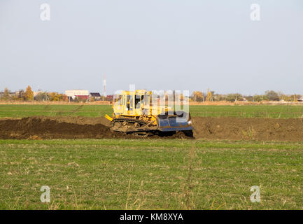 The yellow tractor with attached grederom makes ground leveling. Work on the drainage system in the field. Stock Photo