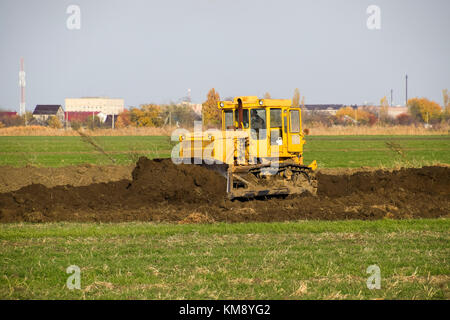 The yellow tractor with attached grederom makes ground leveling. Work on the drainage system in the field. Stock Photo