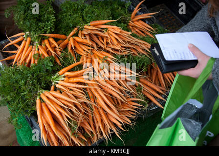 A woman with a shopping list chooses carrots on a vegetable stall at the award winning farmers market in Stroud, Gloucestershire, UK Stock Photo