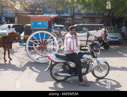 Man sitting on motorcycle and talking on mobile phone, horsedrawn carriage and vehicles parked in background at Mysore, Mysuru, Karnataka, India. Stock Photo