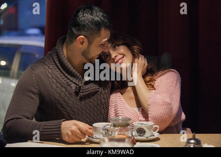 Couple sitting by a window at local coffee place and enjoying winter night with hot drinks and casual conversation. Shot from outside through the wind Stock Photo
