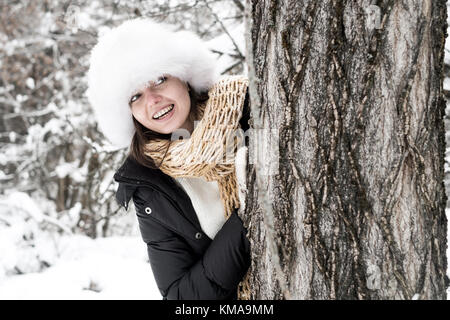 Portrait of smiling young woman wearing winter-clothes standing hide behind tree in forest Stock Photo