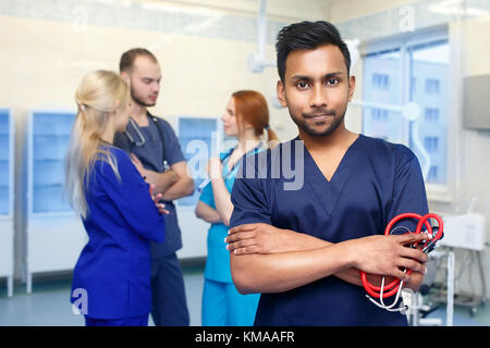Asian male doctor in front of team, looking at camera with medical team in background. Multiracial team of young doctors in a hospital standing in a operating room. Stock Photo