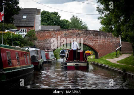Man steers his narrowboat towards a brick bridge whilst another approaches on the Birmingham & Fazeley canal, Staffordshire, England. Stock Photo