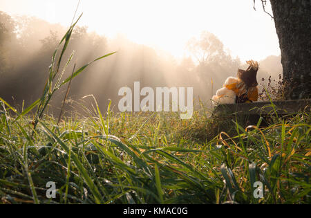 Partnership theme image with a teddy bear and a scarecrow embraced, sitting together on a wooden bench under a tree and enjoying the sun. Stock Photo
