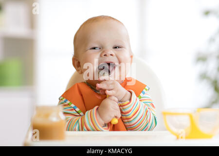 Cheerful baby child eats food itself with spoon. Portrait of happy kid boy in high-chair. Stock Photo