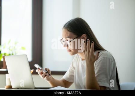 Young furious woman receiving bad negative news, spam in messages on smartphone. Broken mobile cellphone not working and needs charge Stock Photo