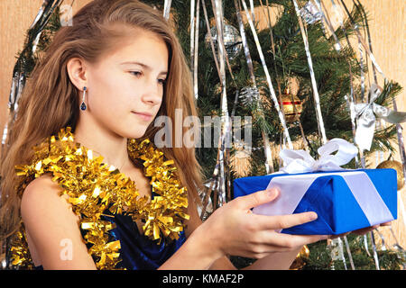 Beautiful teen girl near christmas tree happily looks at gift in her hands Stock Photo