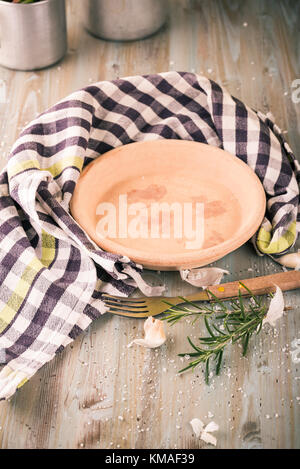 Vertical photo of empty clay plate placed on wooden board with checkered towel arranged around. Parley twig, garlic, salt grains and fork are next to  Stock Photo
