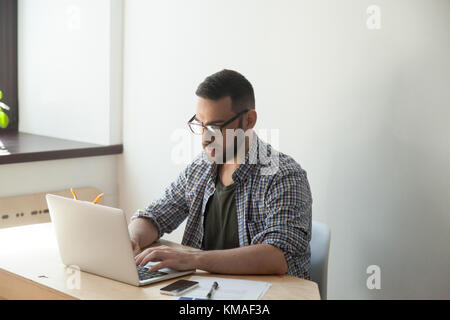 Millennial businessman sitting and typing on laptop in home office. Confident manager wearing glasses analyzing data on computer Stock Photo