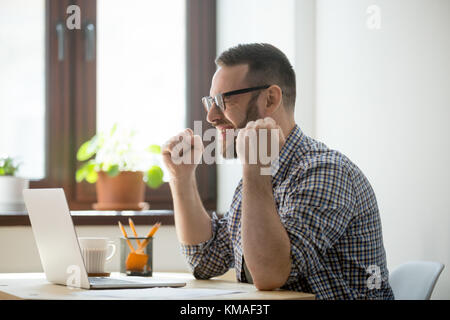 Young happy manager celebrating his victory in job. Millennial bearded worker expressing positivity and raising arms. Success working concept Stock Photo