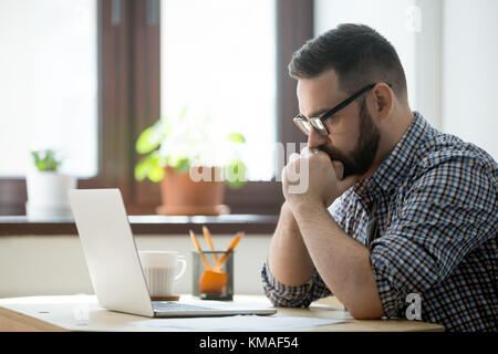 Young bearded manager working and reading data on laptop in home office. Thoughtful casual businessman thinking about job and looking on screen of not Stock Photo