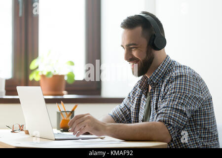 Handsome man with beard listening music in headphones and looking on screen of laptop. Young manager having a rest and playing in computer games or ma Stock Photo
