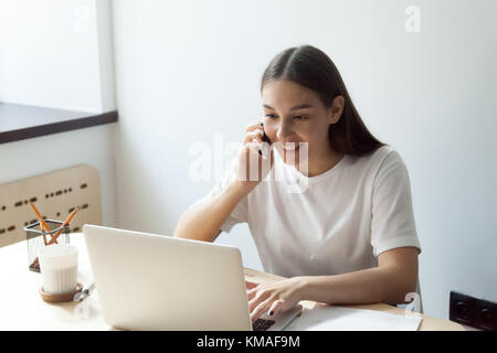 Young happy woman talking on mobile phone and preparing for exams  on laptop. Communicative millennial student chatting in social network Stock Photo