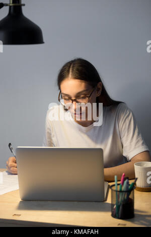 Portrait of young businesswoman sitting at desk and working on laptop in evening. Tired woman in glasses writing and checking documents overtime. Vert Stock Photo