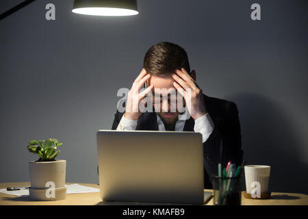 Portrait of tired anxious male worker with headache. Stressed man in glasses holding head in hands, looking at laptop and thinking about deadline. Ove Stock Photo