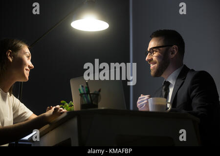 Millennial businesspeople have meeting in office at night. Happy smiling coworkers sitting at desktop and discussing new project under light of lamp i Stock Photo