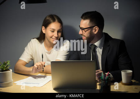Attractive millennial businesswoman looking on laptop and smiling. Young bearded businessman in glasses and suit discussing with colleague new success Stock Photo
