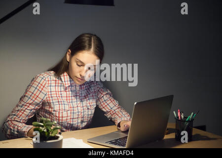 Millennial businesswoman in casual shirt working on laptop in office at night shift. Concentrated female worker checking documents and deals. Stock Photo