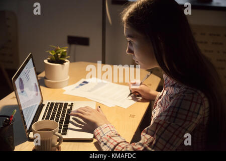 Young serious businesswoman in casual shirt checking work data on laptop and signing signature on business document. Woman employee afraid of deadline Stock Photo