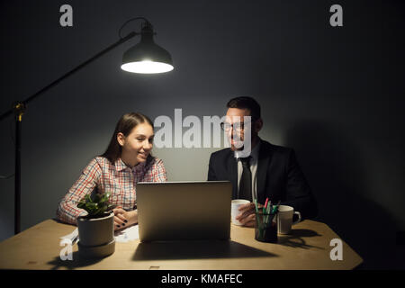 Positive millennial businessman and businesswoman sitting at desktop in office, looking on laptop, planning project and smiling. Night webinar in smal Stock Photo