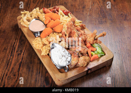 wooden board on a table with assorted fried appetizers: BBQ wings, fried squid rings, fries, grilled cheese, sauce Stock Photo