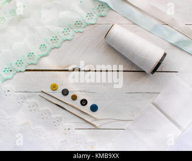 Thread spool, sewing thread line, metal needles and shirt buttons, with frill soft cloth on top of a white wooden background, centered Stock Photo