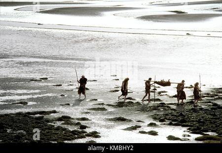 Neolithic prehistoric cave men caveman cavemen cave man walking on mud flat tidal estuary carrying deer food carcass and spears Stock Photo
