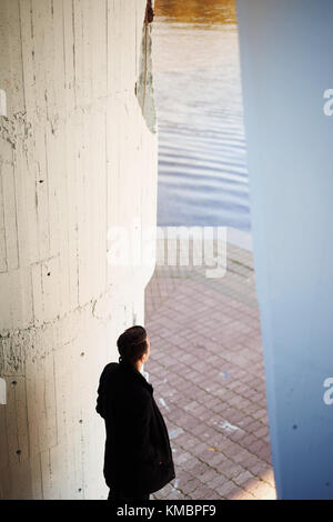 young tall man in a black coat standing at the water and looks into the distance Stock Photo