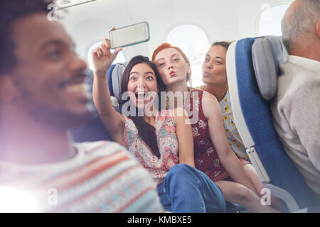 Playful young friends with camera phone taking selfie on airplane Stock Photo