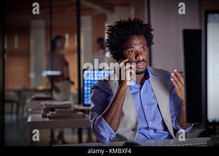 Businessman gesturing,talking on cell phone,working late at computer in dark office Stock Photo