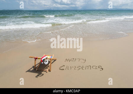 Merry Christmas written in sand write on tropical beach.Deck chair with Santa Claus sunglasses Stock Photo