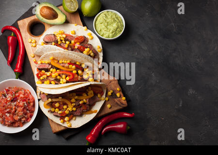 Mexican tacos with marbled beef and vegetables on a dark stone background. Next are the sauces, guacamole and salsa. Top view with copy space. Stock Photo