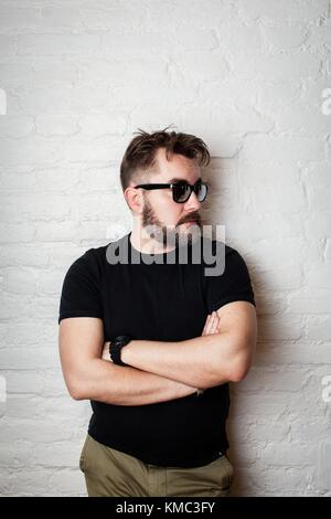 Portrait of a bearded serious man in sunglasses and casual clothes against a white brick wall. Stock Photo