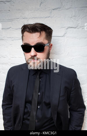 Portrait of a bearded serious man in sunglasses and a business black suit against a white brick wall. Stock Photo