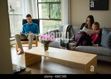Father, mother and daughter reading book in living room Stock Photo